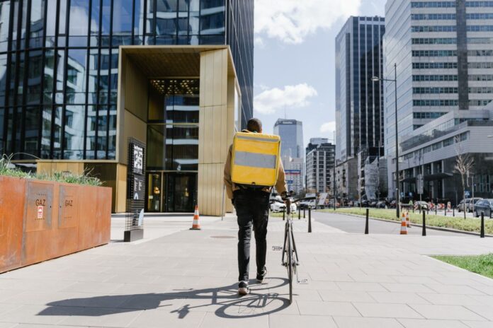 Maaltijdbezorger met gele tas loopt met fiets door zakenwijk, bij artikel over de Wet platformwerk